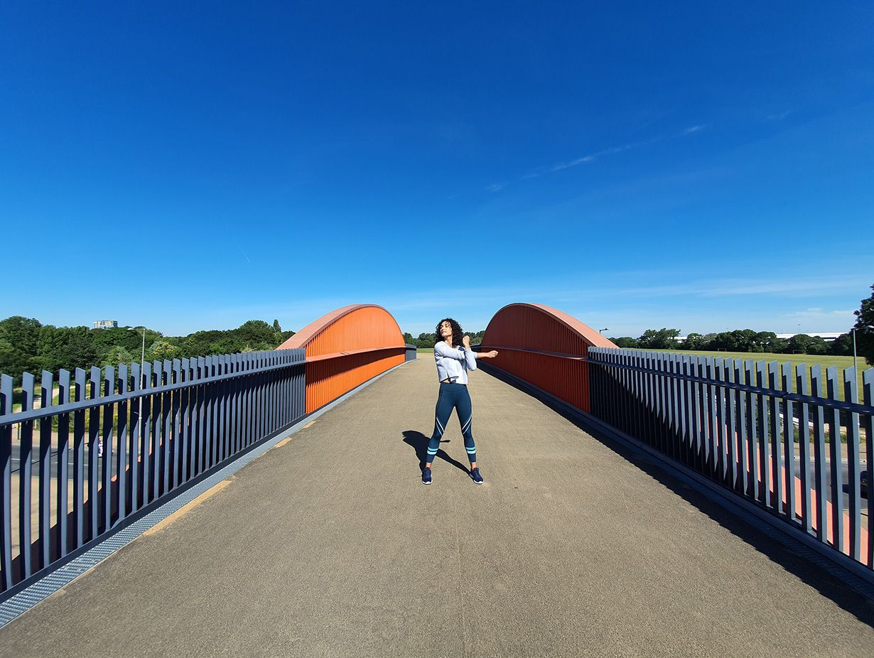 Photo captured by the Ultra-wide Camera of a woman stretching on a bridge with grey gating and orange accents against a bright blue sky