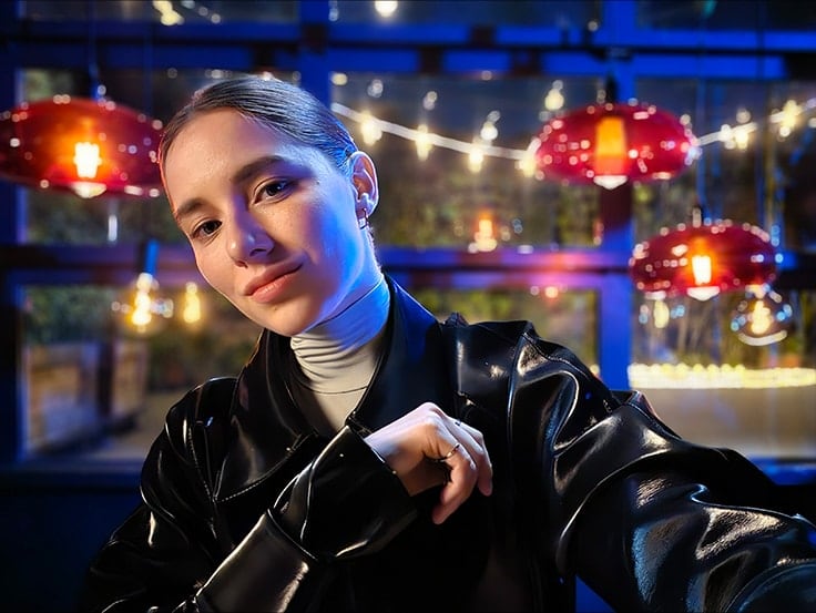 A woman takes a colorful, clear selfie in a dark setting with string lights and lanterns behind …