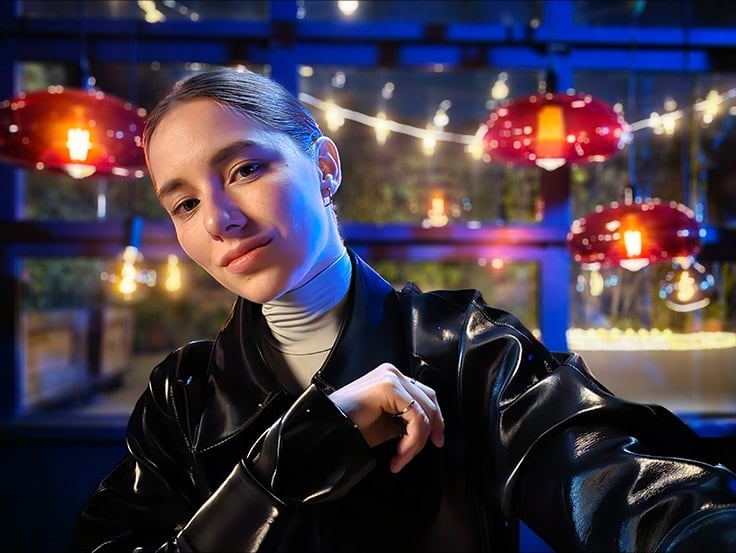 A woman takes a colorful, clear selfie in a dark setting with string lights and lanterns behind her. The photo was taken with Galaxy S23 Ultra using ISO 1600, wide mode, and an aperture of F 2.2.