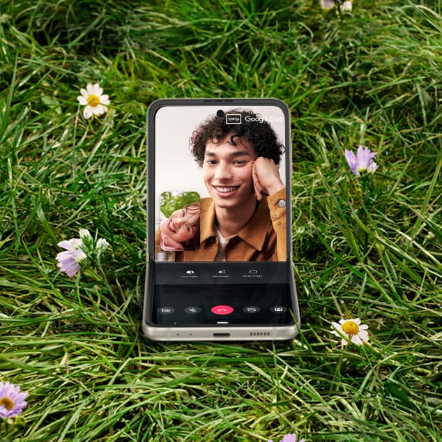 Galaxy Z Flip3 5G in Flex mode in the middle of a field with a video call on screen. The person on the other side of the call is a man holding a drink.